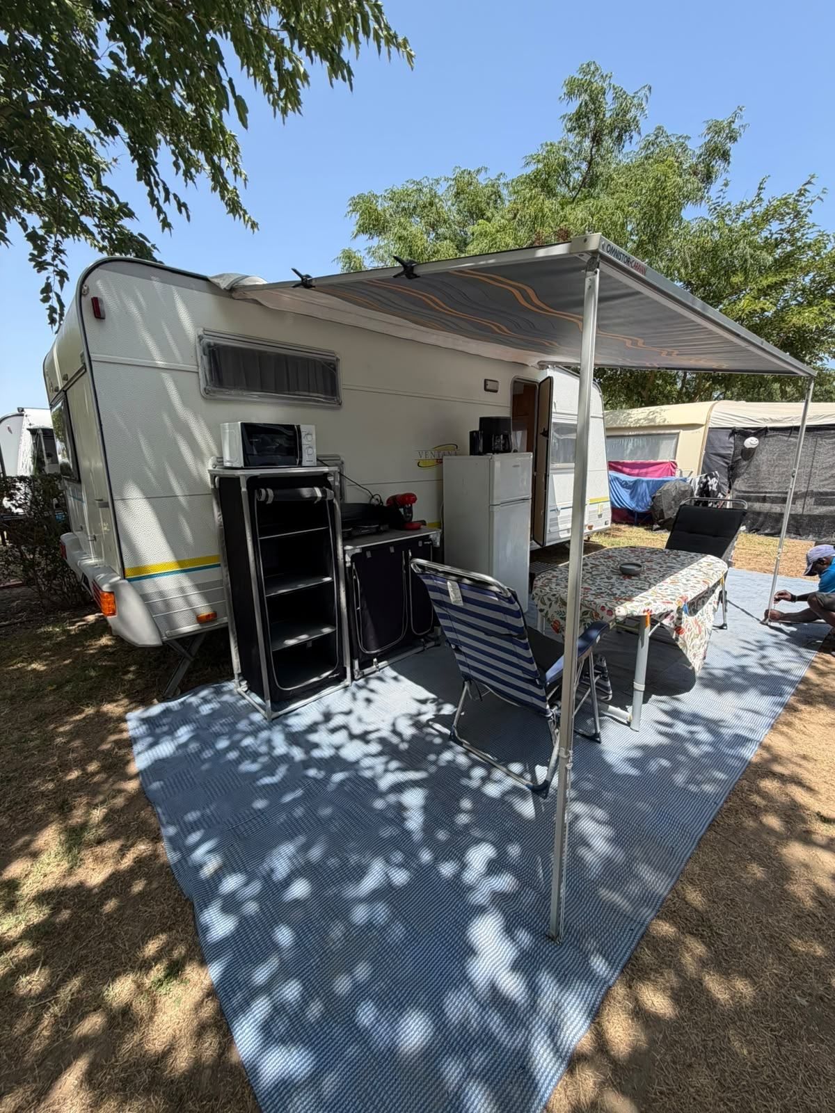 Caravan with a roll-out awning, table, chairs and fridge on a shaded pitch