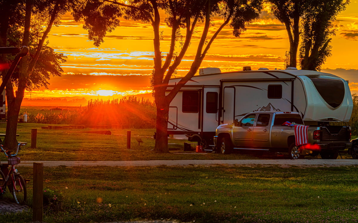 Rv Fifth Wheel and truck parked at campsite with sun setting in background with trees on grass