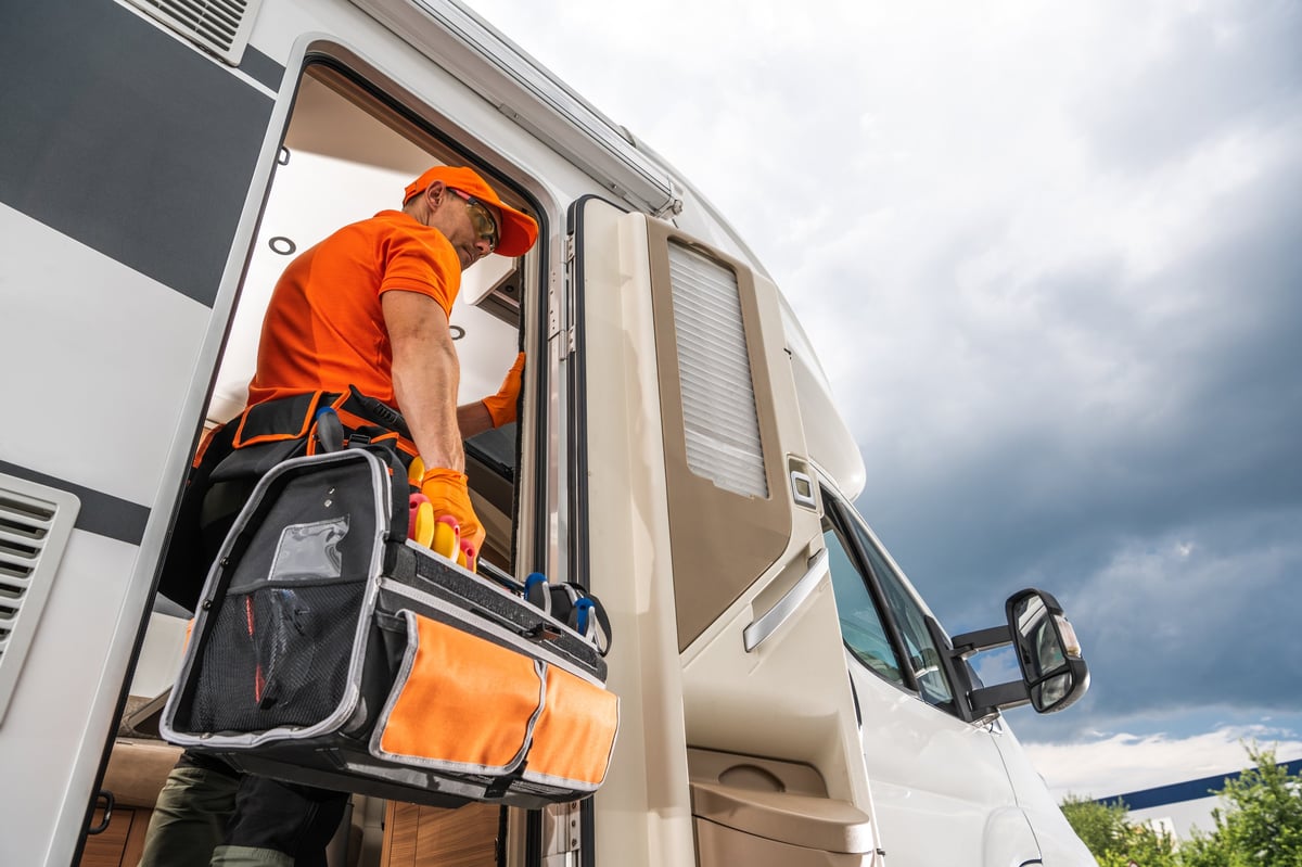 A technician wearing an orange shirt and carrying a tool bag enters an RV
