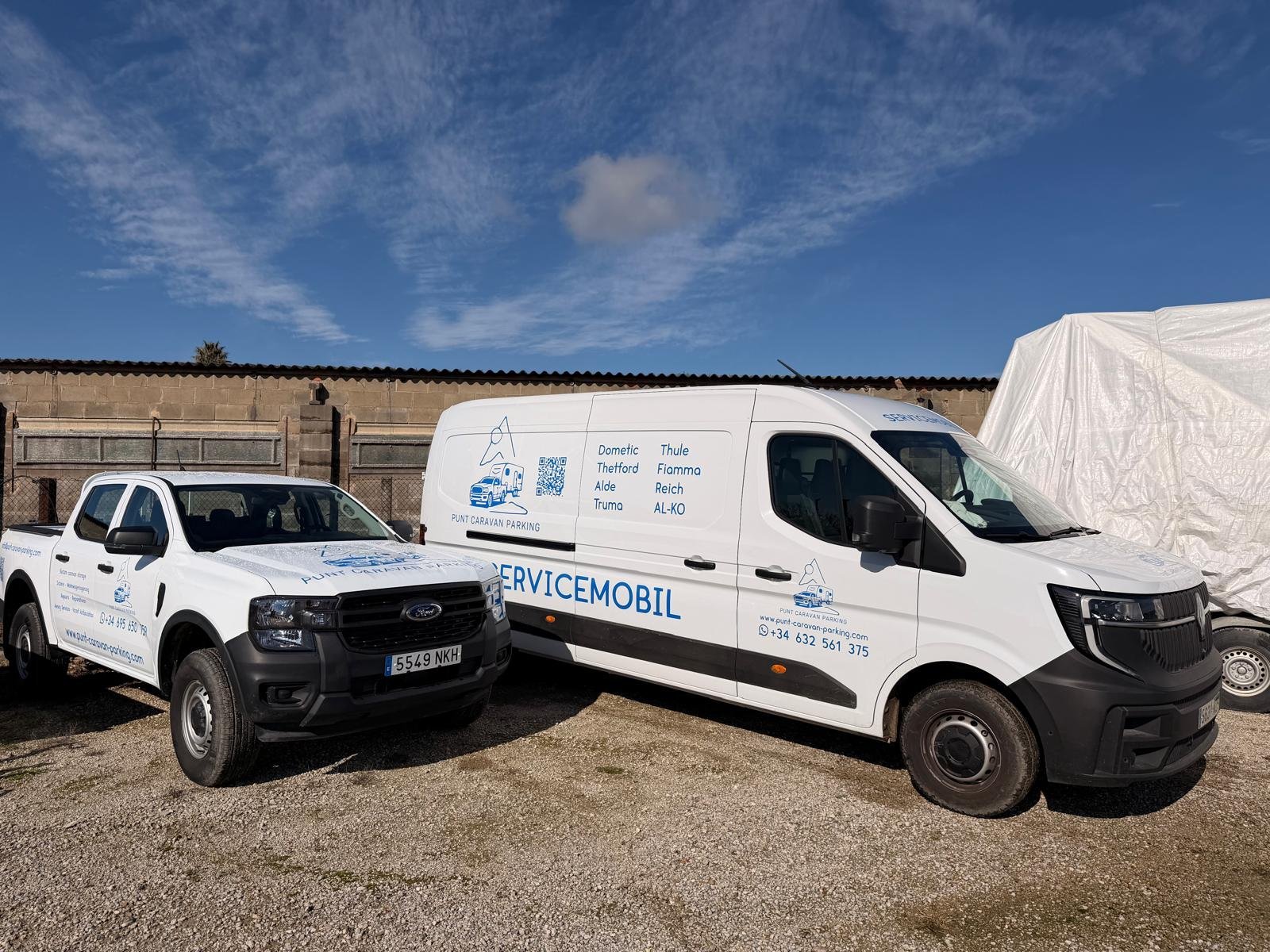 Two white service vehicles parked in an industrial yard with a tarp-covered structure in the background under blue sky